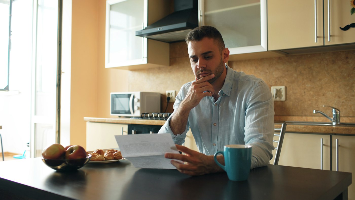 Worried man reading a bill at his kitchen table while taking stock of his debts